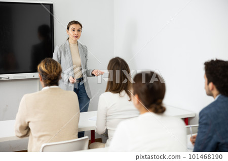Young female business coach explaining topic while standing against group of people sitting at desks in front of her 101468190