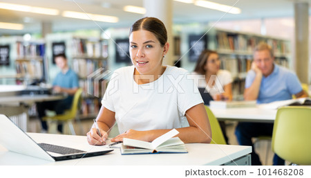 Focused European casual female writing in notepad and using laptop in the library Focused European casual female writing in notepad and using laptop in the library 101468208