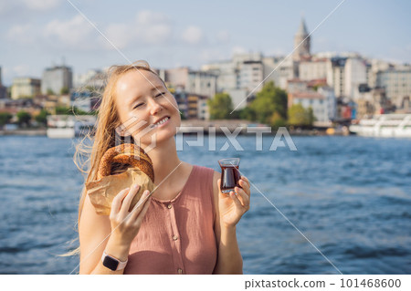 Woman in Istanbul having breakfast with Simit and a glass of Turkish tea. Glass of Turkish tea and bagel Simit against golden horn bay and the Galata Tower in Istanbul, Turkey. Turkiye 101468600