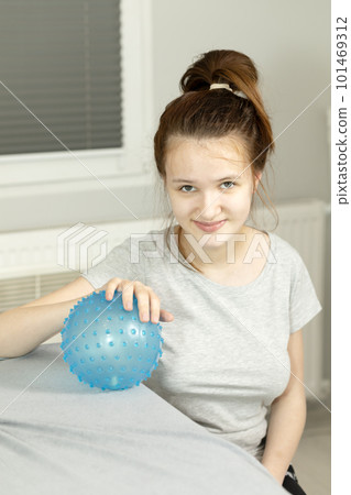 Portrait Of Cheerful Girl With Cerebral Palsy Sitting in Chair, Holding Blue Massage Ball with Pimples in Hospital. Rehabilitation. Balance And Posture Maintenance, Cure. Vertical plane Portrait Of Cheerful Girl With Cerebral Palsy Sitting in Chair, Holding Blue Massage Ball with Pimples in Hospital. Rehabilitation. Balance And Posture Maintenance, Cure. Vertical plane 101469312