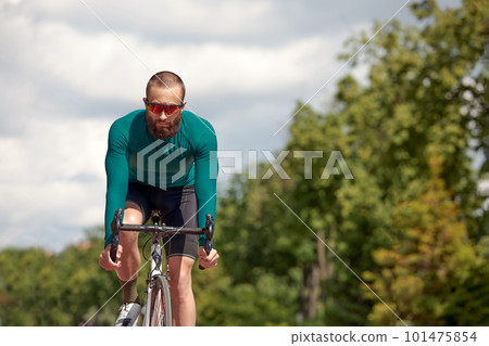 Strong Male cyclist in sportswear, glasses and protective helmet walking with his bike in the forest to take a break after riding. Sky blue in the background. Strong Male cyclist in sportswear, glasses and protective helmet walking with his bike in the forest to take a break after riding. Sky blue in the background. 101475854