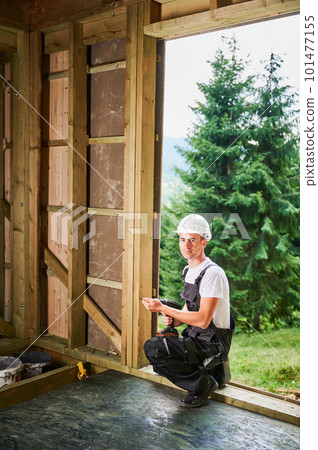 Carpenter constructing wooden framed house. Man worker working with screwdriver, wearing work overalls and helmet. Concept of modern eco-friendly construction. 101477155
