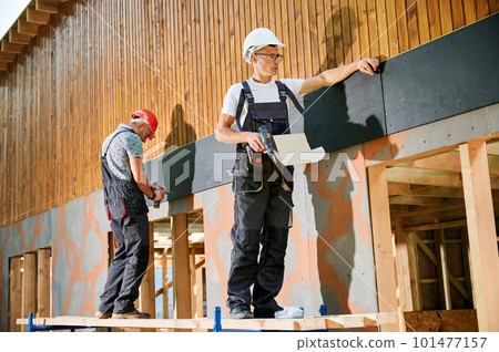 Carpenters constructing wooden framed house. Two men workers cladding facade of house with cement particle boards, fastening them with screwdrivers. Concept of modern eco-friendly construction. 101477157