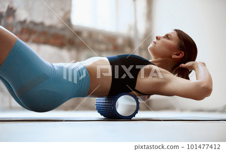 Muscles relaxation. Young girl with sportive body training, doing body massage with foam roller on mat indoors on daytime 101477412