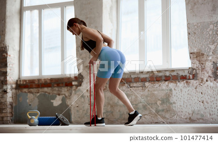 Young girl with sportive slim body training with fitness expanders indoors on daytime. Keeping body fit and strong Young girl with sportive slim body training with fitness expanders indoors on daytime. Keeping body fit and strong 101477414