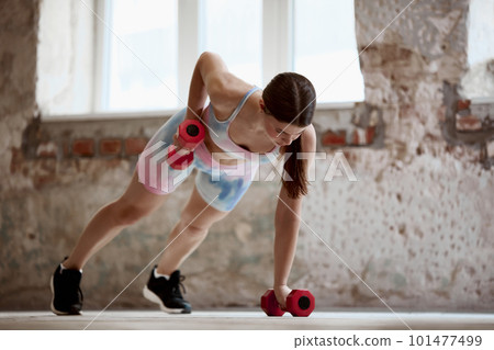 Young sportive girl with slim strong body training, standing in plank position and doing exercises with dumbbells. Press and hands 101477499