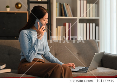 Young adult asian woman having phone conversation and using laptop on couch in cozy living room 101477659