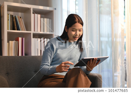 Smiling adult woman sitting on couch browsing wireless internet on digital tablet. Technology, people and lifestyle concept 101477685