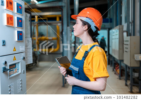 Boiler room. Portrait of a young female worker in helmet and uniform with a tablet in her hand. Side view. The concept of industrial production Boiler room. Portrait of a young female worker in helmet and uniform with a tablet in her hand. Side view. The concept of industrial production 101482282