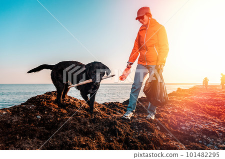 A young Caucasian female volunteer in a jacket, with a garbage bag in her hands, gives a plastic bottle to her dog. In the background, the sea and the sky. The concept of environmental protection 101482295