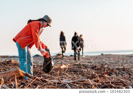 A Caucasian woman in a jacket with a garbage bag in her hands is cleaning up on the beach after a storm. In the background, the sea, the sky, and other cleaners. Concept of environmental pollution A Caucasian woman in a jacket with a garbage bag in her hands is cleaning up on the beach after a storm. In the background, the sea, the sky, and other cleaners. Concept of environmental pollution 101482303