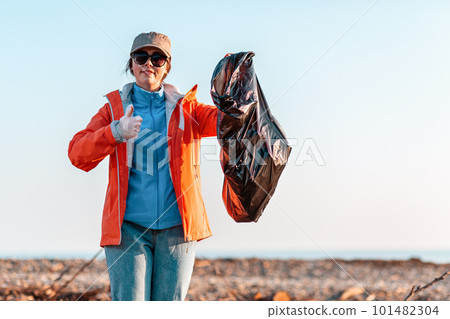 A young Caucasian woman in a jacket, holding a garbage bag and pointing at it with her finger. In the background, the beach after the storm, the sea and the sky A young Caucasian woman in a jacket, holding a garbage bag and pointing at it with her finger. In the background, the beach after the storm, the sea and the sky 101482304