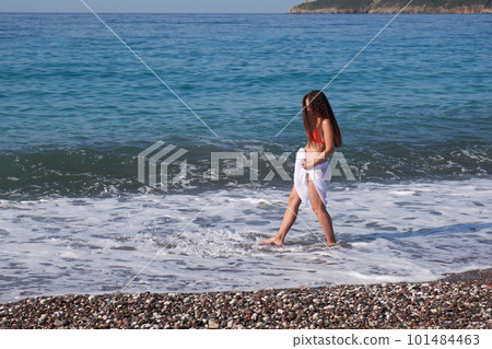 Young brunette woman with long hair in a white pareo and hat walks along the beach. Young brunette woman with long hair in a white pareo and hat walks along the beach. 101484463