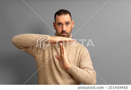 Bearded Hispanic man in his 40s wearing a beige turtleneck making the time-out gesture while looking seriously at the camera, isolated on gray studio background. Bearded Hispanic man in his 40s wearing a beige turtleneck making the time-out gesture while looking seriously at the camera, isolated on gray studio background. 101489488