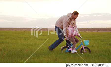 father rides cheerful child two-wheeled bicycle green grass sunset. happy family park. parent rides happy child daughter bicycle. kid girl child with dad park. chidhood dream. happy family concept father rides cheerful child two-wheeled bicycle green grass sunset. happy family park. parent rides happy child daughter bicycle. kid girl child with dad park. chidhood dream. happy family concept 101489538