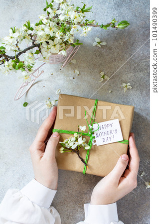 Mother's day or womens day background. Blooming cherry branch and a present in female hands with a congratulatory inscription on a gray stone table. Top view flat lay. Mother's day or womens day background. Blooming cherry branch and a present in female hands with a congratulatory inscription on a gray stone table. Top view flat lay. 101490399