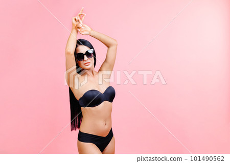 Young woman in summer black swimsuit and sunglasses standing on pink background in studio 101490562