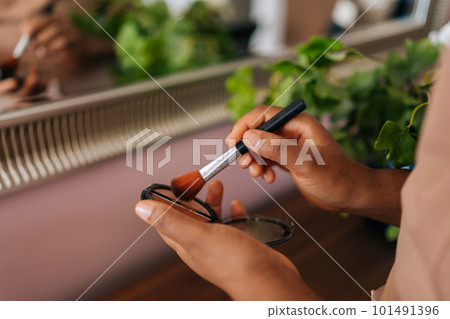 Close-up hands of unrecognizable African American woman holding contour palette and brush standing by mirror in home interior. Beautiful black female putting daily makeup, applying powder indoors Close-up hands of unrecognizable African American woman holding contour palette and brush standing by mirror in home interior. Beautiful black female putting daily makeup, applying powder indoors 101491396