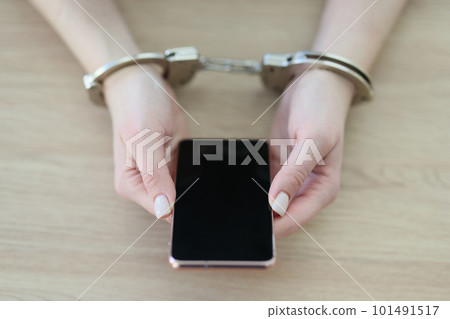 Woman in handcuffs holds modern smartphone sitting at table Woman in handcuffs holds modern smartphone sitting at table 101491517