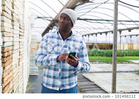 Male gardener standing at greenhouse with smartphone 101498112