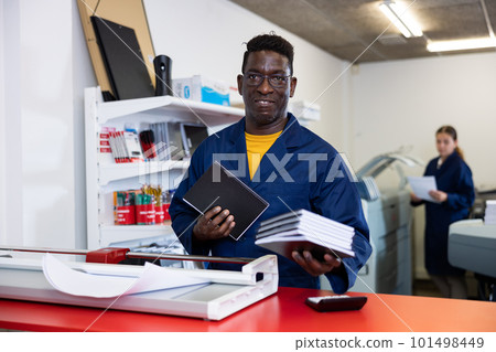 Middle-aged African American man in blue robe uniform smiling at the camera and holding new planners in printer house 101498449