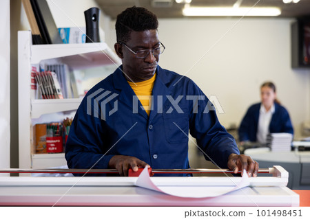 Focused African American man in blue robe uniform trimming the printed work on the cutter in the typography 101498451