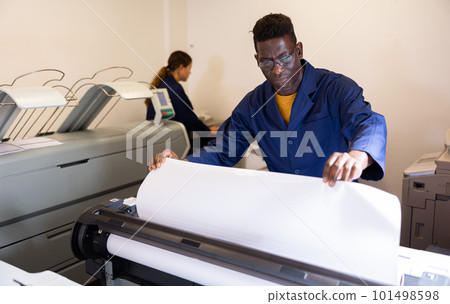 Serious middle-aged African American man in uniform loading large format paper in a plotter in the print shop 101498598