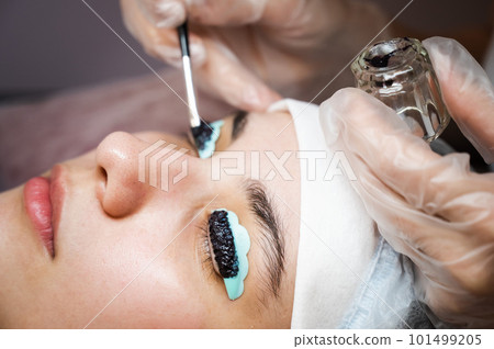 Close-up portrait of a woman on eyelash lamination procedure. The master applies tint to the eyelashes.  101499205