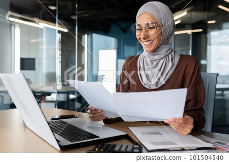 Successful muslim woman in hijab working inside office with documents, joyful financial accountant happy with reports checking financial data and smiling, businesswoman at workplace with laptop. Successful muslim woman in hijab working inside office with documents, joyful financial accountant happy with reports checking financial data and smiling, businesswoman at workplace with laptop. 101504540