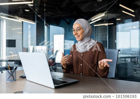 Successful and happy woman inside office at workplace, businesswoman boss in hijab talking on video call using laptop sitting at table muslim woman joyfully gesturing to colleagues in online meeting 101506193