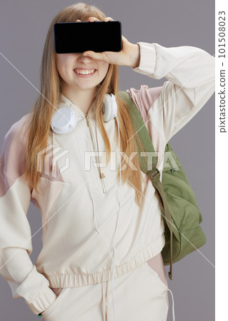 happy girl in beige tracksuit showing smartphone blank screen 101508023