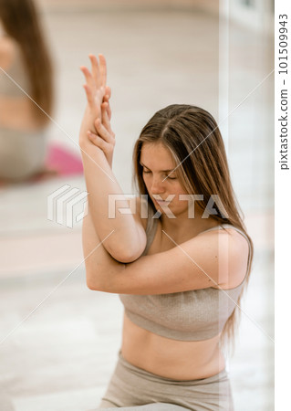 Young woman doing yoga in the gym. A girl with long hair and in a beige tracksuit stands in a cow pose on a pink carpet. A woman performs Gomukhasana. 101509943