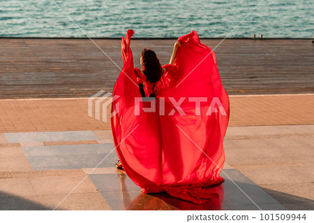 Sunrise red dress. A woman in a long red dress against the backdrop of sunrise, bright golden light of the sun's rays. The concept of femininity, harmony. 101509944