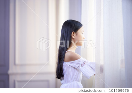 beautiful little asian girl with long black hair looking out of window in bedroom 101509976