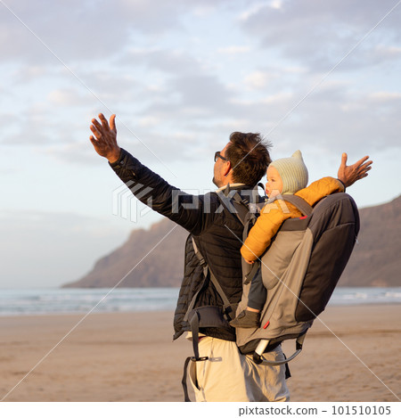 Father rising hands to the sky while enjoying pure nature carrying his infant baby boy son in backpack on windy sandy beach of Famara, Lanzarote island, Spain at sunset. Family travel concept Father rising hands to the sky while enjoying pure nature carrying his infant baby boy son in backpack on windy sandy beach of Famara, Lanzarote island, Spain at sunset. Family travel concept 101510105