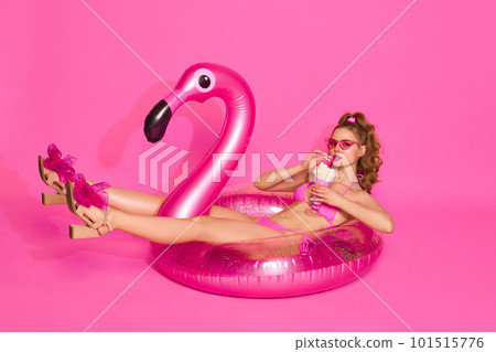 Portrait of blond girl wearing swimsuit drinking milkshake and laying on inflatable flamingo over pink studio background. Pool party 101515776