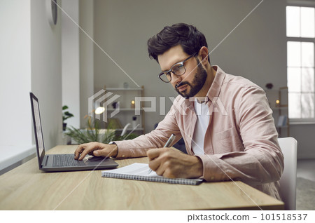 Man sitting at his desk at home, studying online, using his laptop and taking notes 101518537