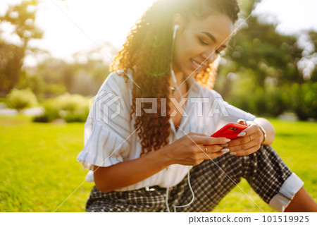 Young woman sitting on green grass in park with phone. Pretty woman listening to music. 101519925