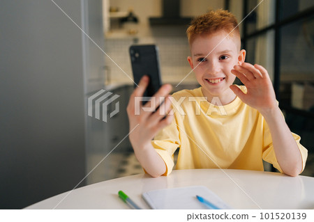 Portrait of happy child schoolboy waving hand, looking at smartphone distance learning class using mobile application, watching online lesson, video calling app. Homeschooling with modern tech usage 101520139