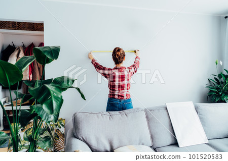 Young woman in plaid shirt doing measuring with a measure tape on the wall. Girl wants to put a picture on the wall at home. Housekeeping work. Doing repair herself. DIY, equality in work concept. 101520583