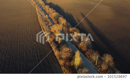 Beautiful landscape view old asphalt road with trees and shadows between large plowed agricultural fields of black soil on sunny autumn evening. Flying over dug agrarian fields of earth. Aerial drone Beautiful landscape view old asphalt road with trees and shadows between large plowed agricultural fields of black soil on sunny autumn evening. Flying over dug agrarian fields of earth. Aerial drone 101528697