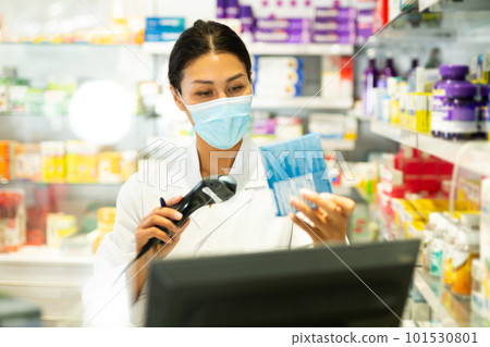 Portrait of female pharmacist in medical mask working at the cash register in pharmacy - scans barcode on medicine package Portrait of female pharmacist in medical mask working at the cash register in pharmacy - scans barcode on medicine package 101530801