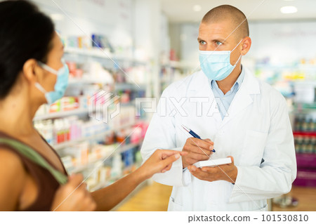 Pharmacist in protective mask writes in notebook the name of the drugs that buyer tells him to 101530810