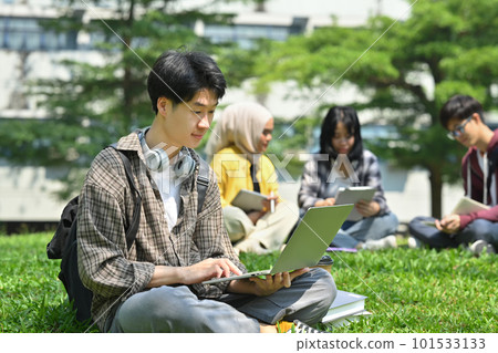 Young asian man student using laptop on green grass in front of university building. Education, technology and lifestyle concept 101533133