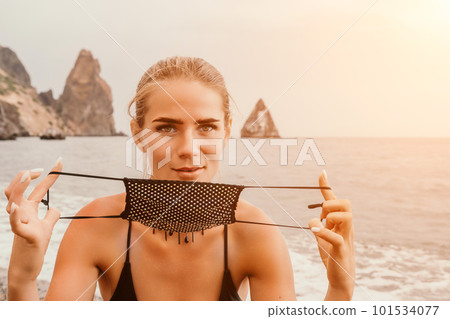 Woman travel portrait. Happy woman with long hair poses on a red volcanic rock at the beach. Close up portrait cute woman in black bikini, smiles at the camera, with the sea in the background. Woman travel portrait. Happy woman with long hair poses on a red volcanic rock at the beach. Close up portrait cute woman in black bikini, smiles at the camera, with the sea in the background. 101534077