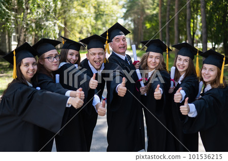 A group of graduates in robes give a thumbs up outdoors. Elderly student. A group of graduates in robes give a thumbs up outdoors. Elderly student. 101536215