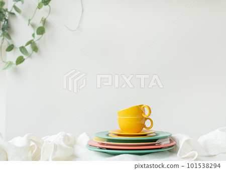Empty yellow cups and brightly colored plates on a white table covered with a linen tablecloth, a flower pot on the wall in the background. Preparation for serving. 101538294
