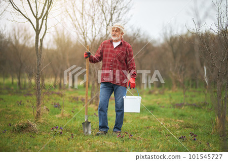 Front view of old man working in orchard in spring. Male gardener in panama hat and gloves standing, looking, holding spade and bucket. Concept of countryside and agriculture. 101545727