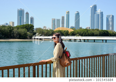 The skyline of Sharjah is visible from the waterfront - a woman tourist with a backpack enjoys the view of the water and skyscrapers. Cityscape of Sharjah UAE 101545884
