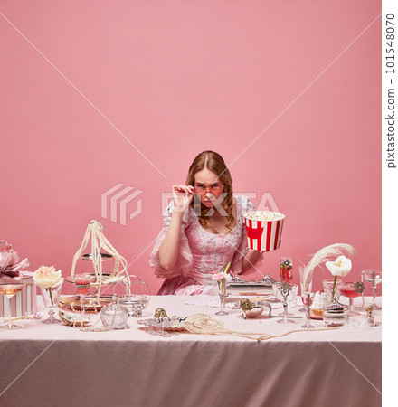 Fairy tale blond princess wearing renaissance dress sitting near the laid table with popcorn on pink studio background. Watching movie with snacks 101548070